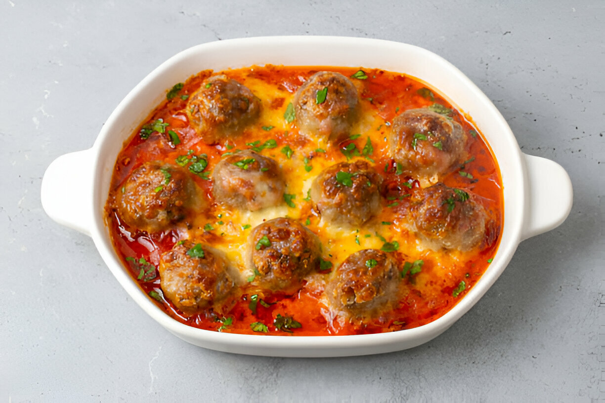 A white serving dish filled with meatballs in a red sauce topped with melted cheese and garnished with green herbs.