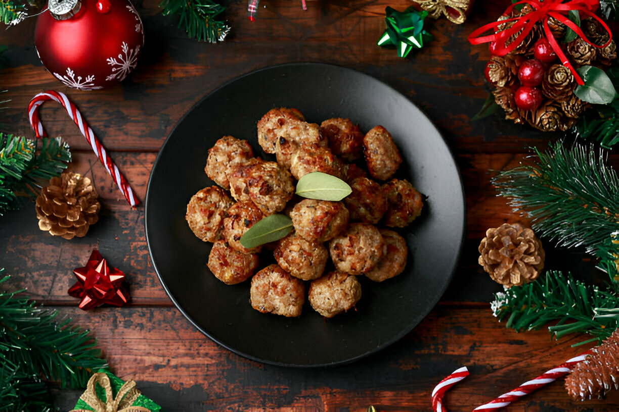 A black plate filled with meatballs garnished with bay leaves, surrounded by Christmas decorations like pine cones, candies, and colorful stars.