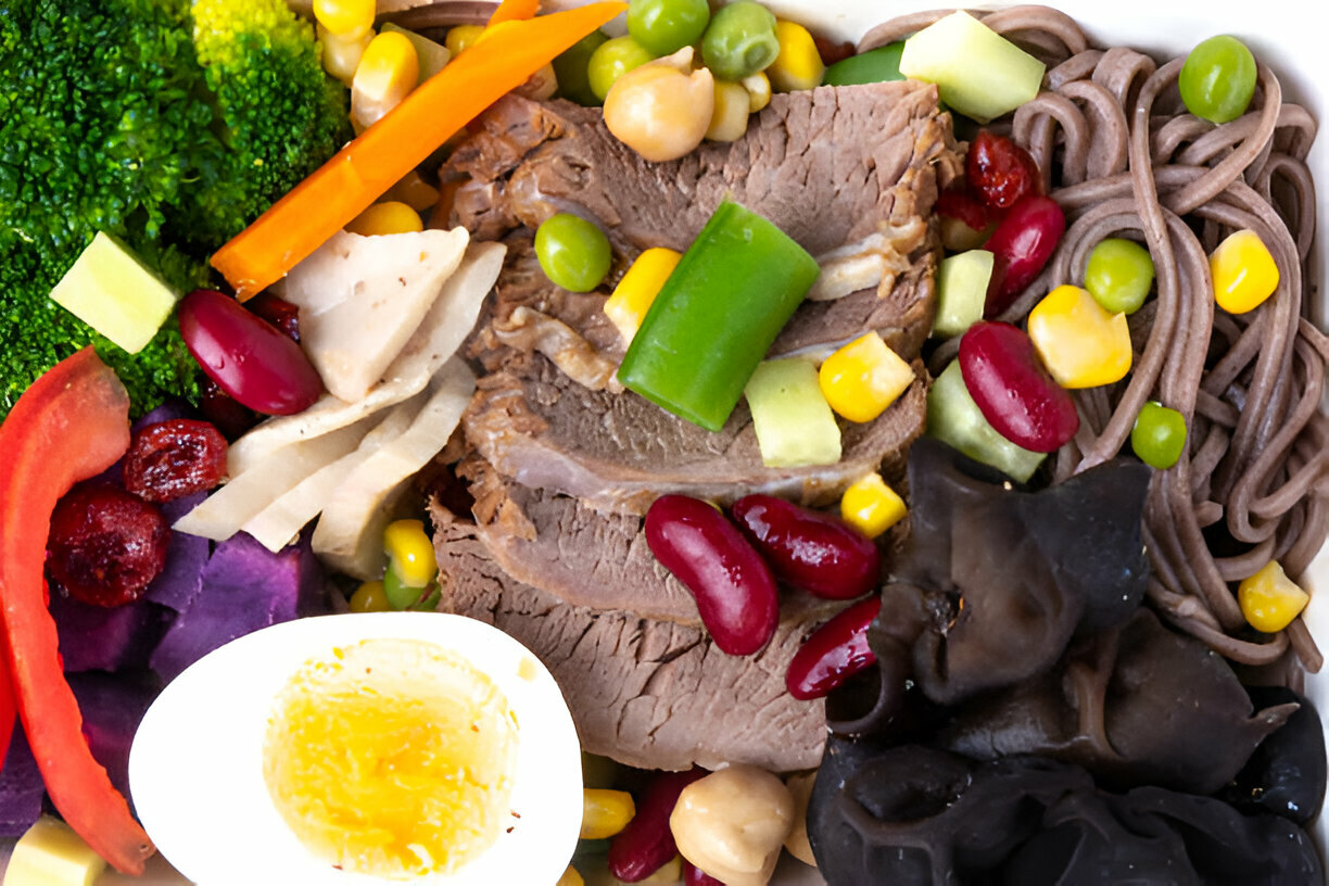 A vibrant bowl of mixed ingredients including broccoli, assorted vegetables, beans, sliced meat, soba noodles, and a soft-boiled egg.