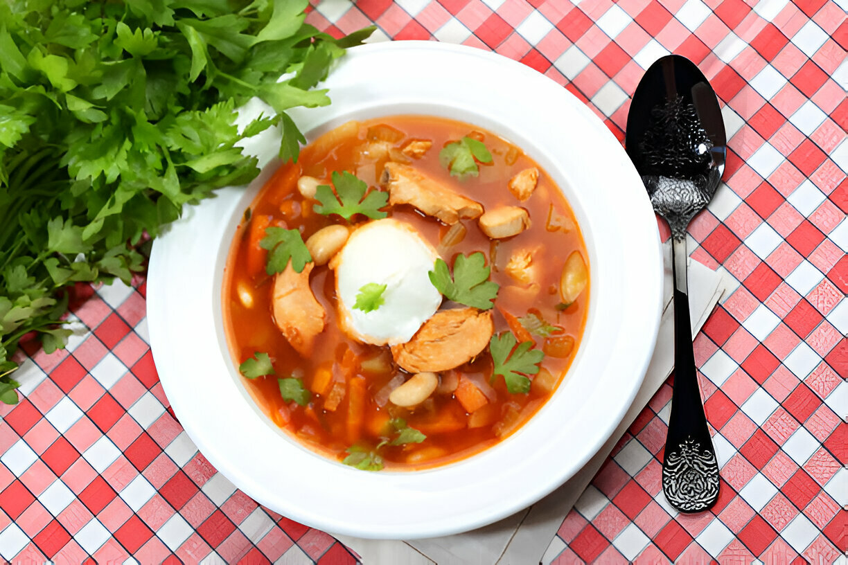 A bowl of flavorful chicken soup topped with parsley and a dollop of sour cream, surrounded by fresh cilantro on a checkered tablecloth.