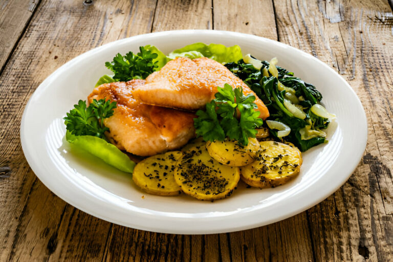 A plate of grilled salmon with herbs, accompanied by sautéed greens and seasoned potatoes on a wooden table.