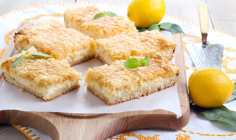 A freshly baked lemon dessert cut into squares on a wooden platter, garnished with mint leaves and surrounded by lemons.