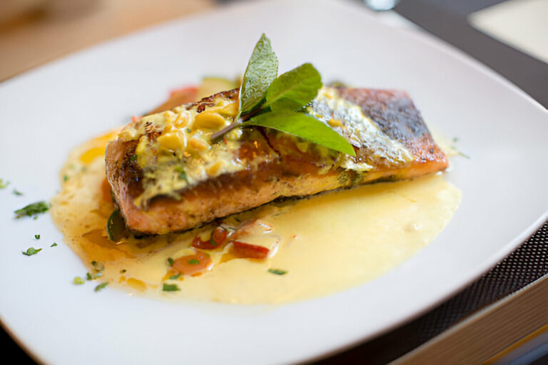 A beautifully plated salmon fillet topped with herbs and garnished with leaves on a white plate.