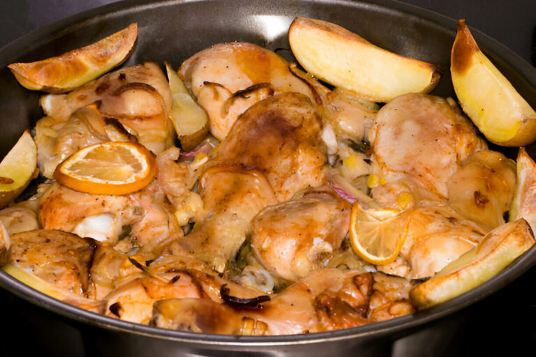 A close-up of roasted chicken pieces with lemons and potato wedges in a dark dish.