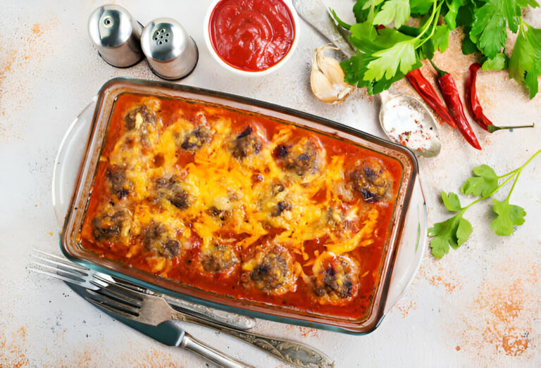 Baked meatballs with cheese in a glass dish, surrounded by fresh ingredients like parsley, garlic, and chili peppers.