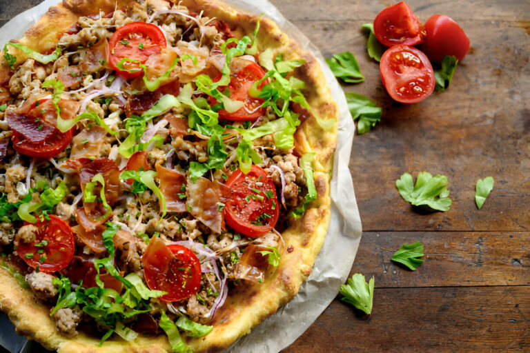 A freshly baked pizza topped with sliced tomatoes, greens, and various meats on a wooden table.