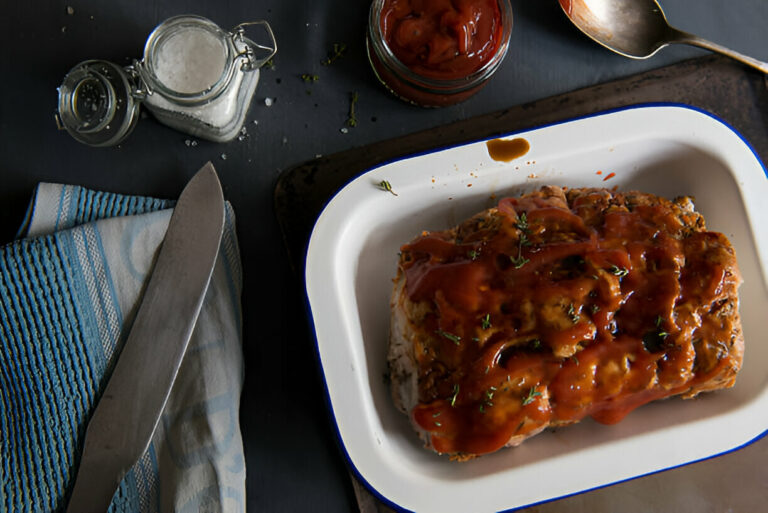 A plate of meatloaf topped with sauce and garnished with herbs, alongside a knife and salt shakers.