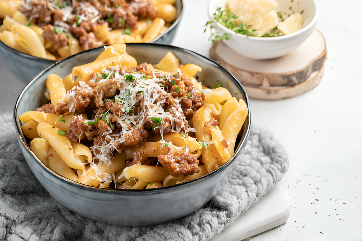 A bowl of pasta with meat sauce, topped with grated cheese and parsley, served alongside a small bowl of cheese on a wooden tray.