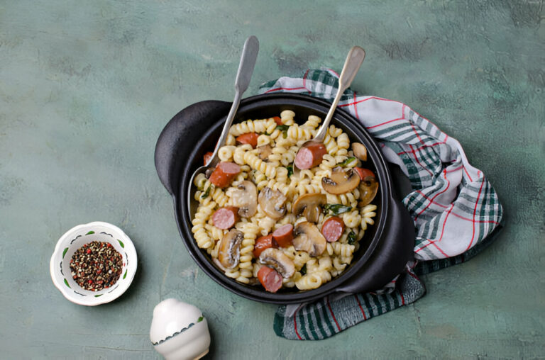 A delicious serving of spiral pasta mixed with sausage, mushrooms, and green vegetables in a black dish, accompanied by a small bowl of mixed pepper seasoning.