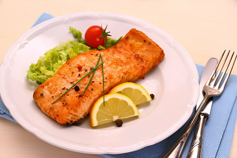 A beautifully plated salmon fillet with lemon slices and a side of lettuce and cherry tomato.