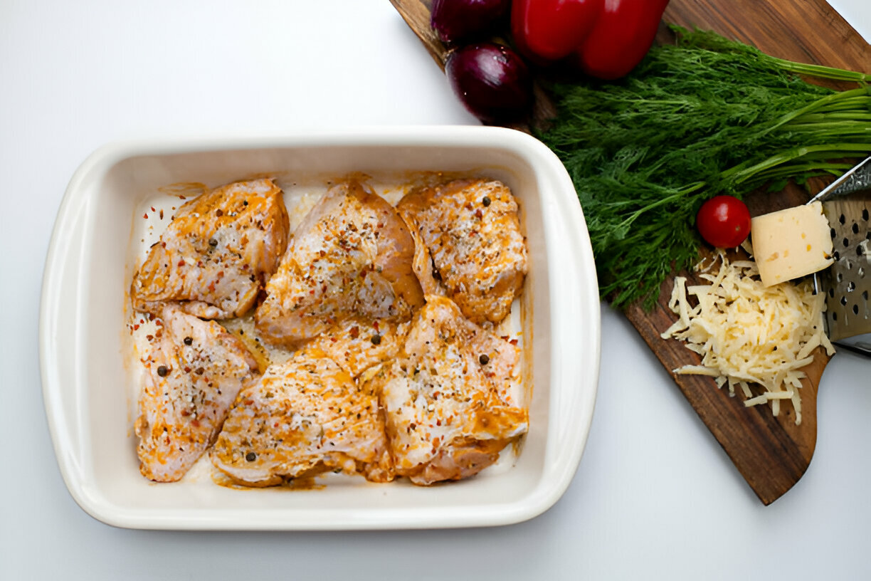 Marinated chicken thighs in a white baking dish, surrounded by fresh vegetables and grated cheese.