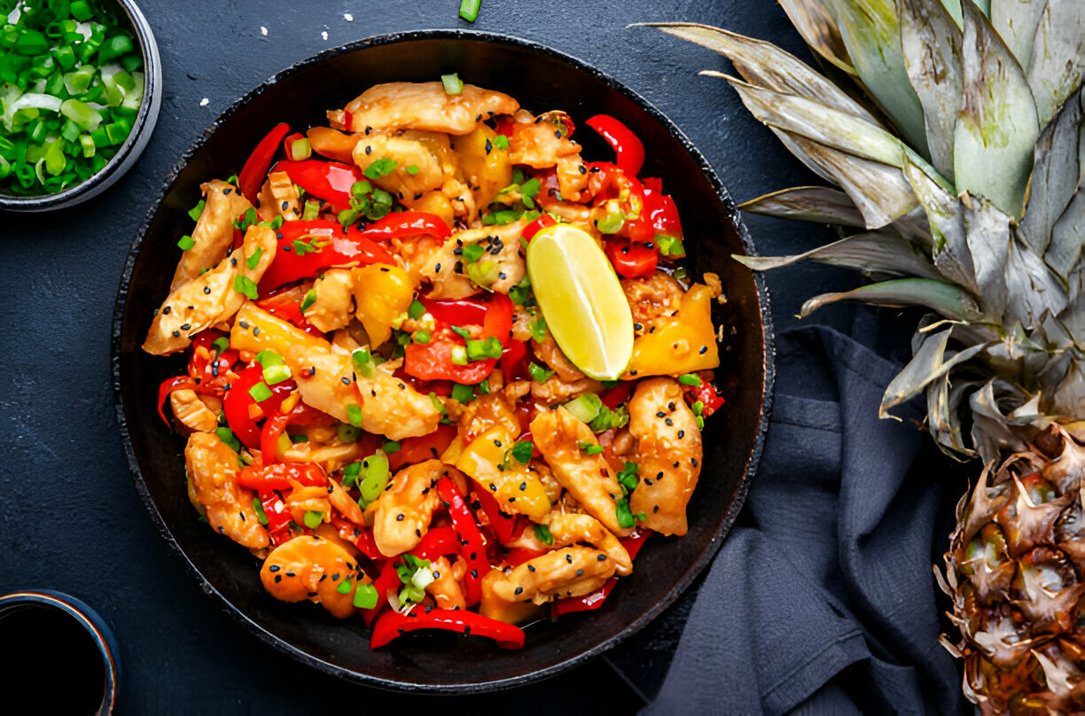 A vibrant bowl of chicken stir-fry with bell peppers, garnished with green onions and a slice of lime, accompanied by a pineapple.