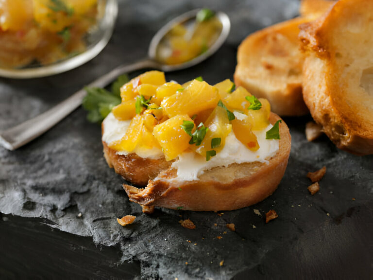 A slice of bread topped with cream cheese and mango salsa, garnished with herbs on a rustic black stone surface.