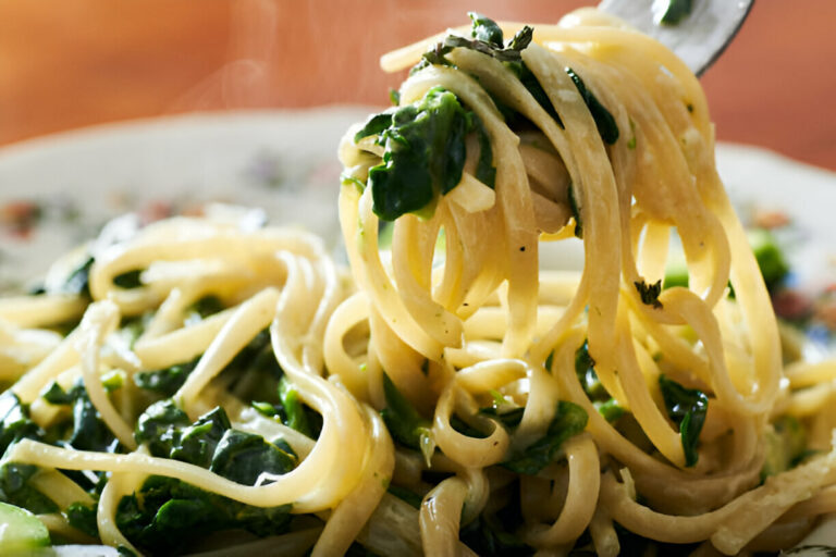 A fork picking up fettuccine pasta mixed with green spinach on a ceramic plate.