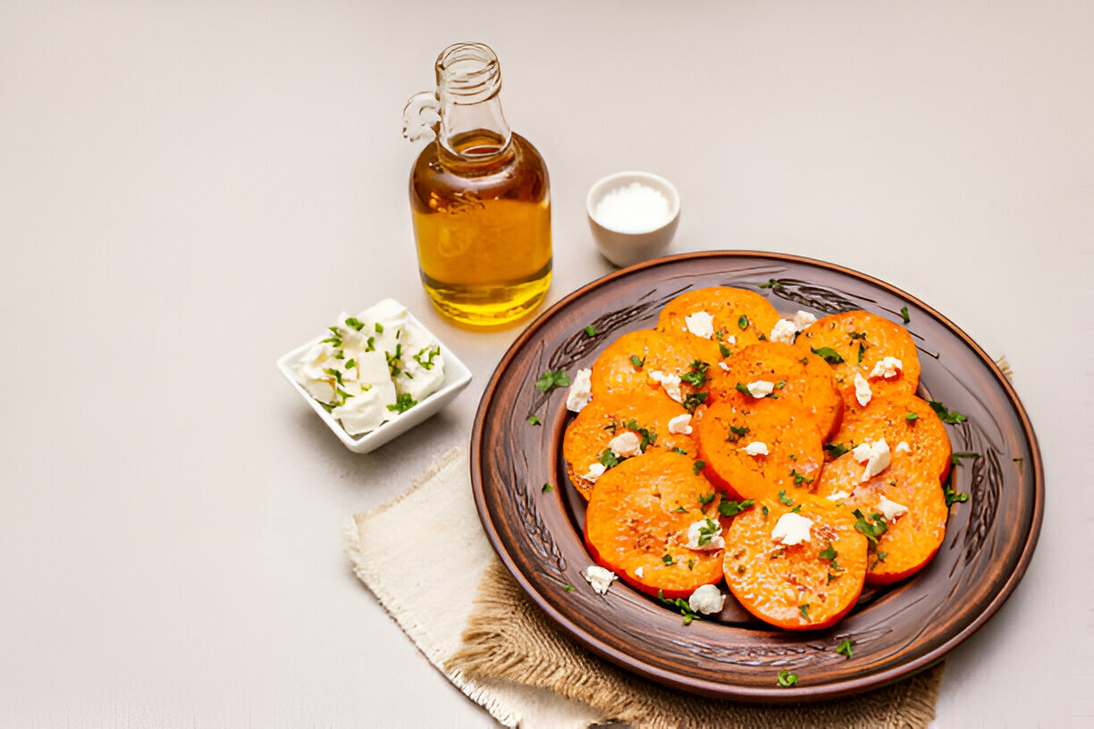 A plate of sliced, roasted orange vegetables topped with crumbled cheese and herbs, accompanied by olive oil and a small dish of white sauce.