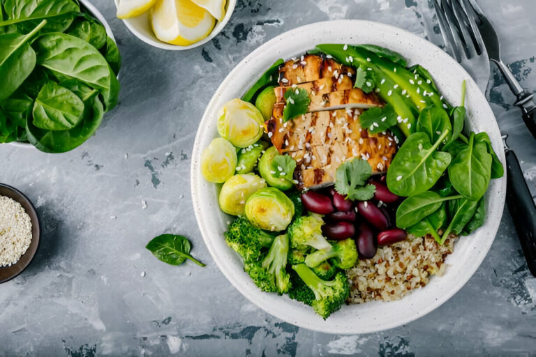 A healthy bowl filled with grilled chicken, colorful vegetables including Brussels sprouts, broccoli, and spinach, served over quinoa, with lemon slices in the background.