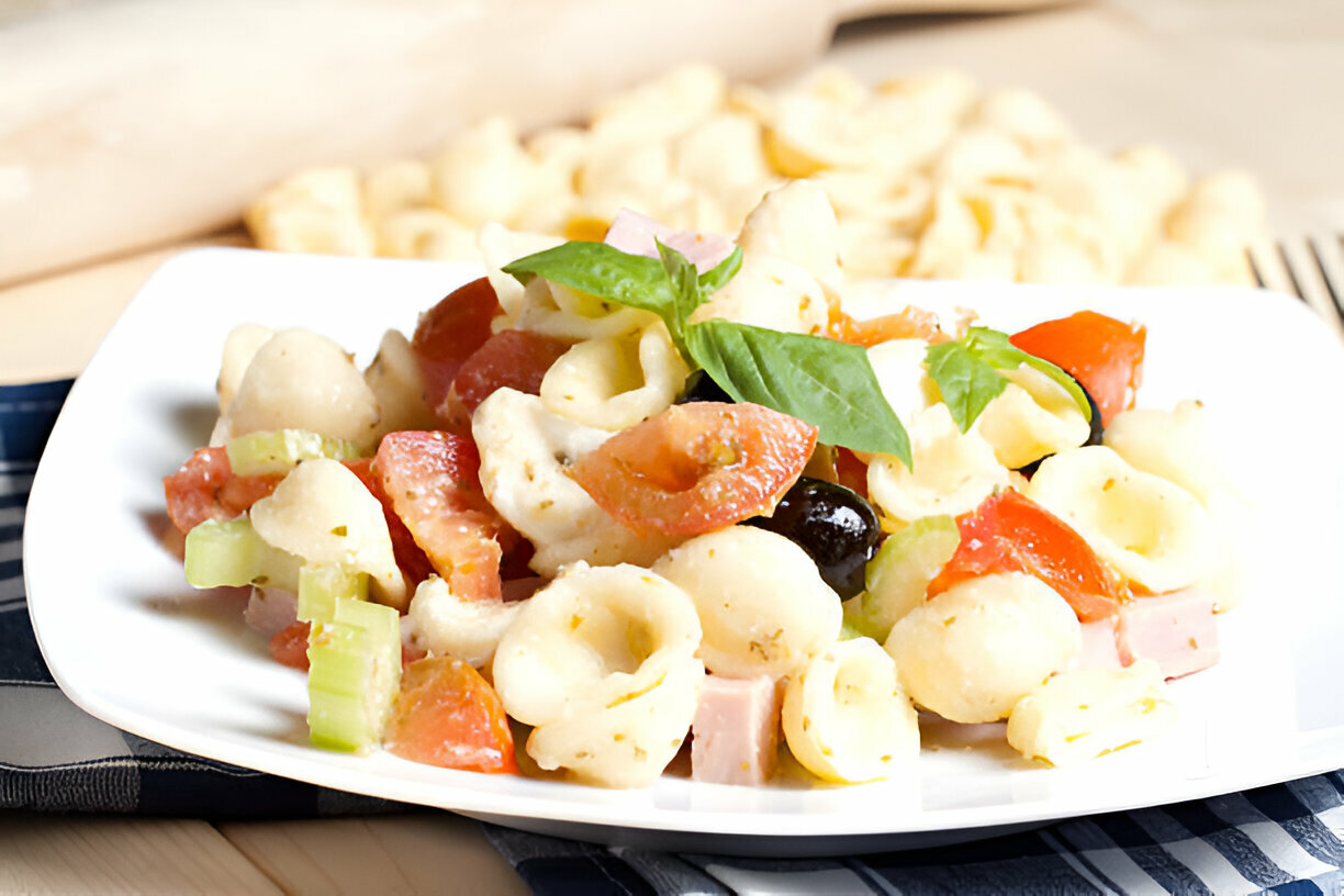 A plate of pasta salad with tomatoes, olives, celery, and basil leaves