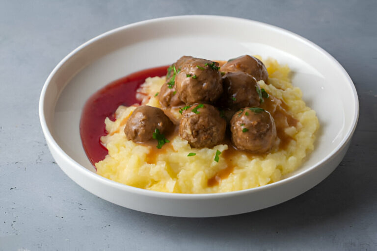 A plate of meatballs served over creamy mashed potatoes with brown gravy and a drizzle of red sauce.