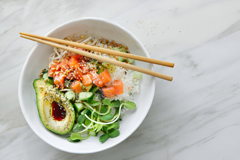 Bowl of fresh sushi ingredients including rice, avocado, cucumber, salmon, and sprouts with chopsticks on top.