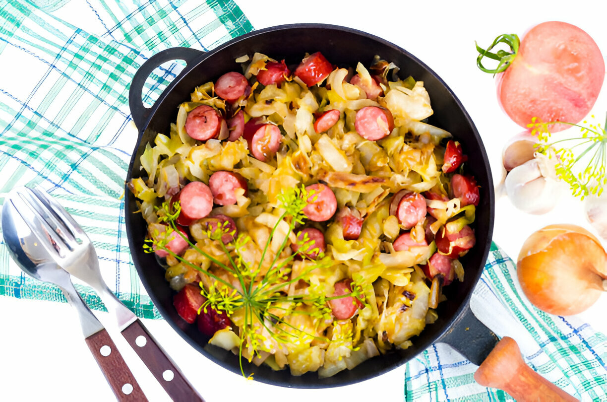 A skillet filled with cooked cabbage and sausage pieces, garnished with fresh herbs, set against a white background with green and blue checkered cloth.