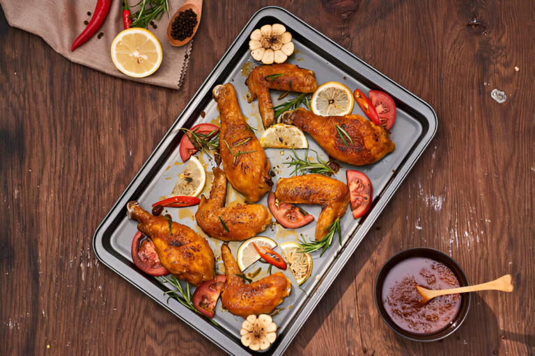 A tray of roasted chicken pieces garnished with lemon slices, tomatoes, and herbs, alongside a bowl of dipping sauce.