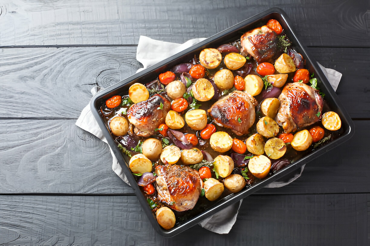 A baking tray filled with roasted chicken thighs, baby potatoes, cherry tomatoes, and herbs.