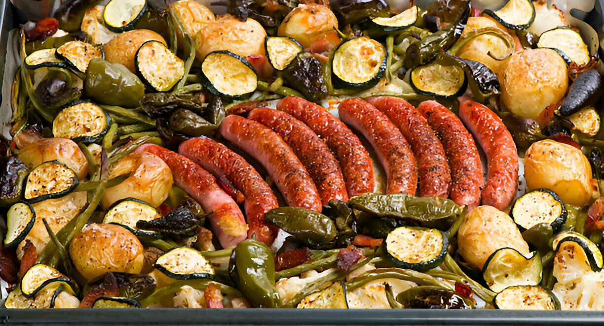 A tray of baked sausages surrounded by various roasted vegetables including zucchini, potatoes, and green peppers.