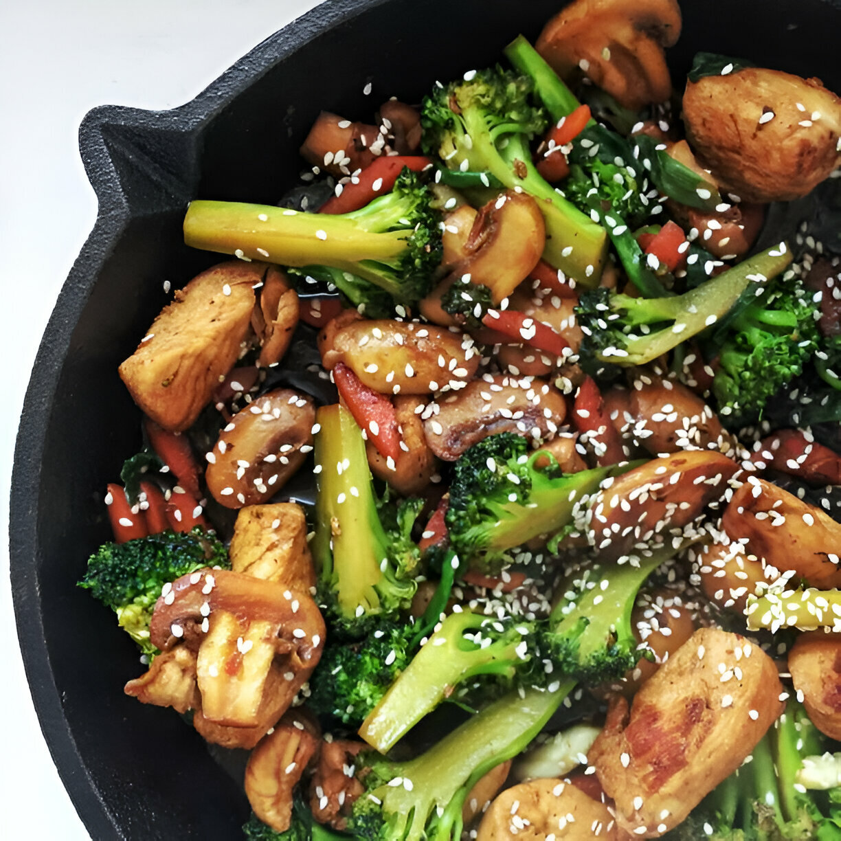 A close-up of a cast iron skillet filled with stir-fried chicken, mushrooms, broccoli, and red peppers, topped with sesame seeds.