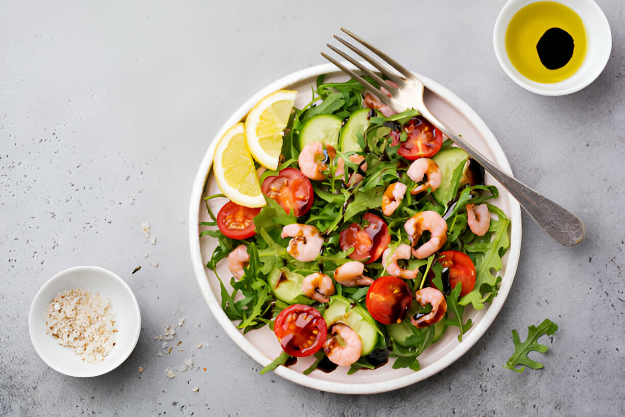 A fresh salad with shrimp, cherry tomatoes, cucumber, and lemon slices served in a white bowl with a fork and olive oil nearby.