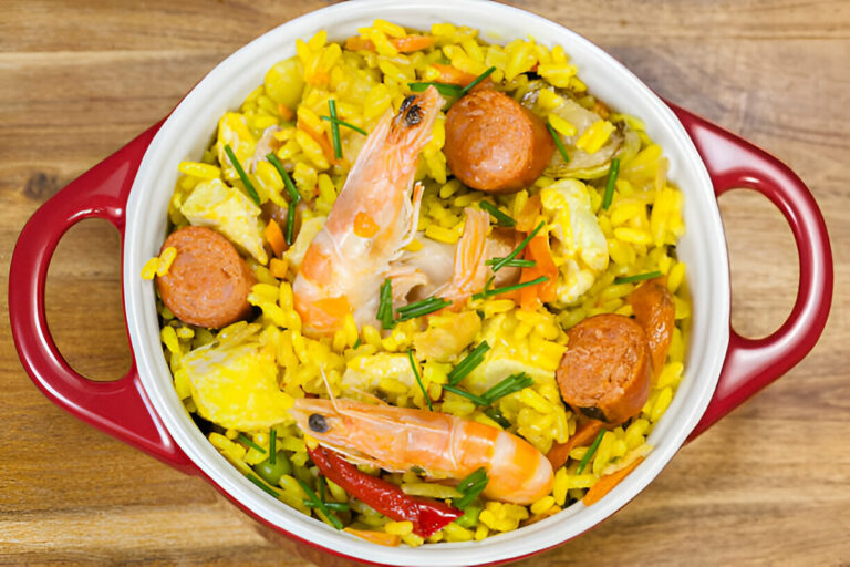 A vibrant dish of yellow rice with shrimp, sausages, and vegetables in a red casserole dish