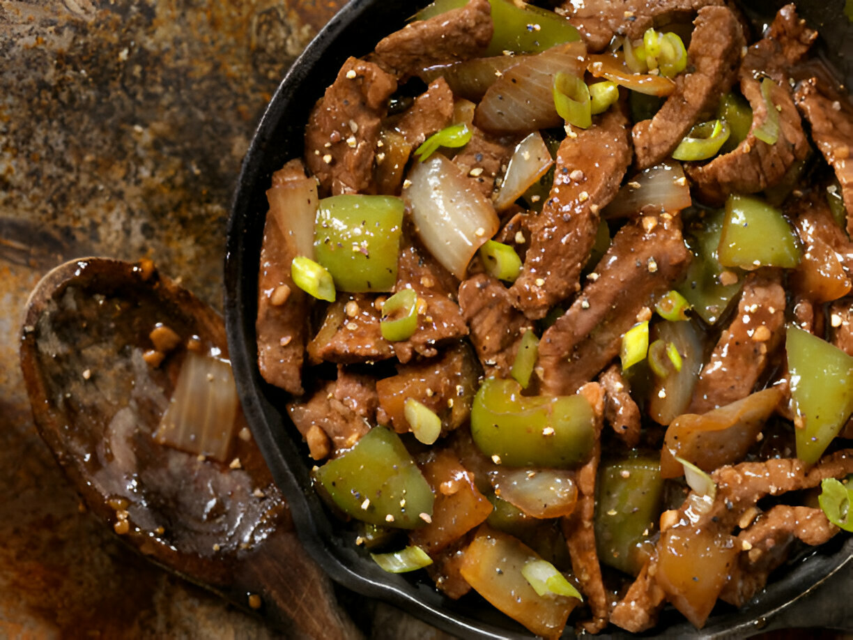 A skillet filled with stir-fried beef, green bell peppers, and onions, garnished with sesame seeds and chopped green onions.