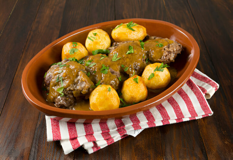 A dish of meat with gravy and round potatoes presented in a brown serving bowl on a striped cloth.