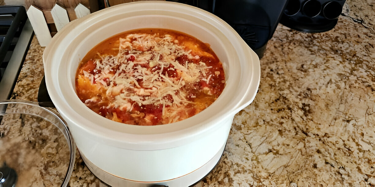 A slow cooker containing a meal with a rich red sauce and cheese topping on a countertop.