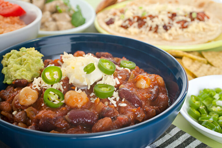 A bowl of hearty chili topped with cheese, sour cream, and sliced jalapeños, accompanied by guacamole, tortilla chips, green onions, and other side dishes.