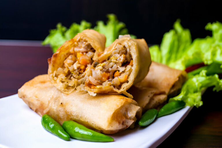 A plate of crispy vegetable spring rolls with one cut in half, revealing a filling of mixed vegetables, served with green chili and lettuce.