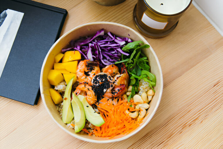 A vibrant bowl of healthy food featuring shrimp, mango, purple cabbage, greens, and quinoa, placed on a wooden table.