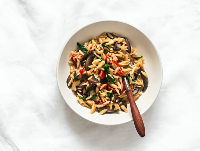 A bowl of orzo pasta with vegetables, garnished with herbs, on a light background.