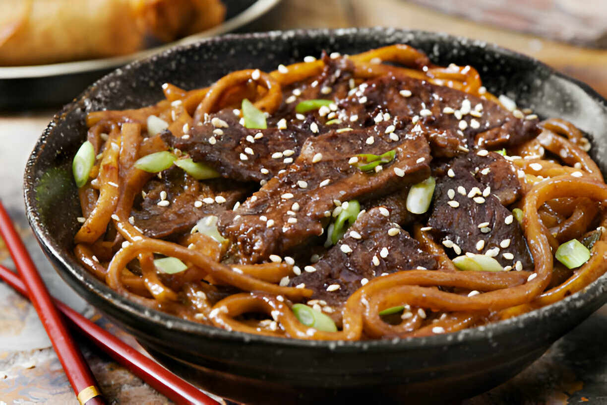 A close-up of a bowl of noodles topped with sliced beef, green onions, and sesame seeds, with chopsticks beside it.