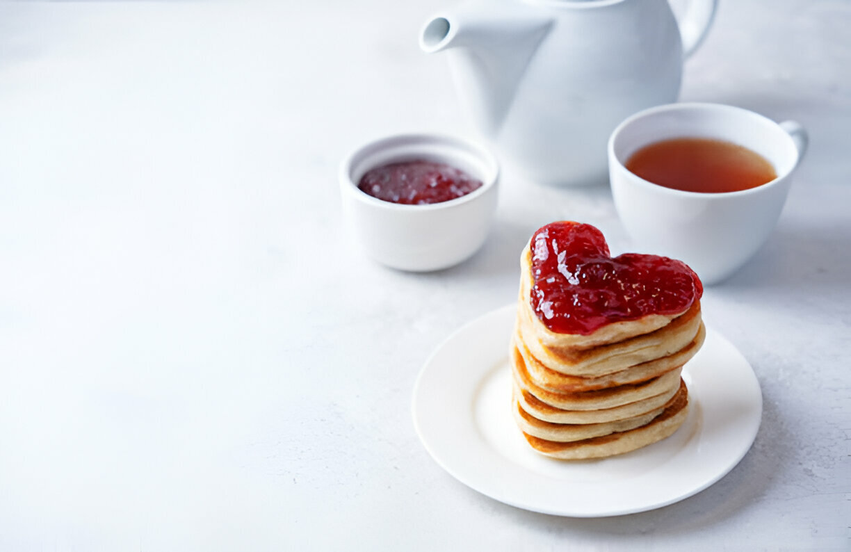 A stack of heart-shaped pancakes topped with red jam, accompanied by a small bowl of jam and a cup of tea.