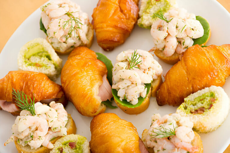 A colorful platter of assorted appetizers featuring croissant-style baked goods topped with shrimp and green garnishes, alongside sushi rolls and other seafood bites.