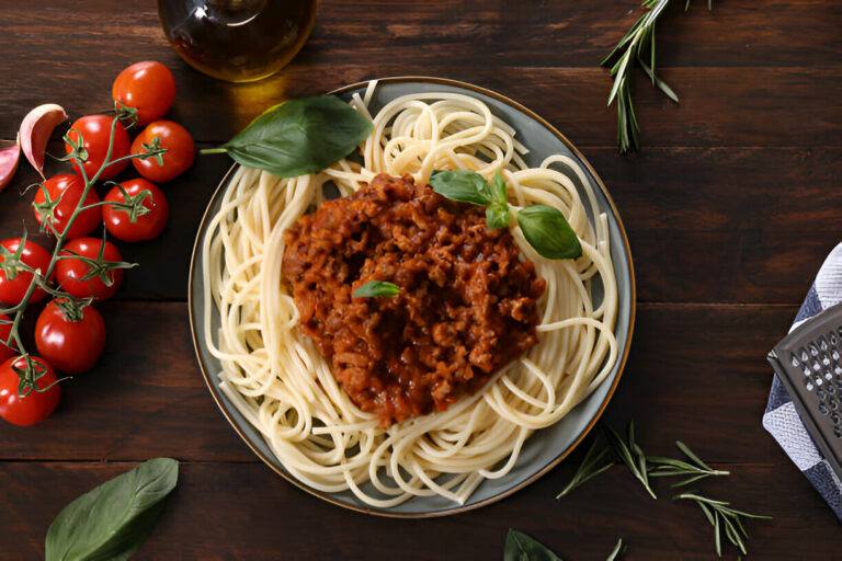 A plate of spaghetti topped with a rich meat sauce, garnished with basil leaves, surrounded by fresh tomatoes and olive oil.