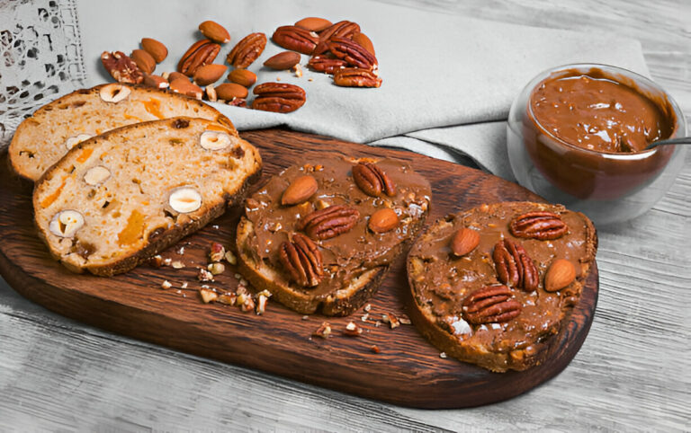 Sliced bread topped with nut butter and nuts on a wooden platter, accompanied by a bowl of nut spread.