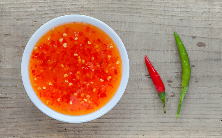 A bowl of chili sauce with chopped red chilies, placed on a wooden surface, accompanied by a red and a green chili pepper.