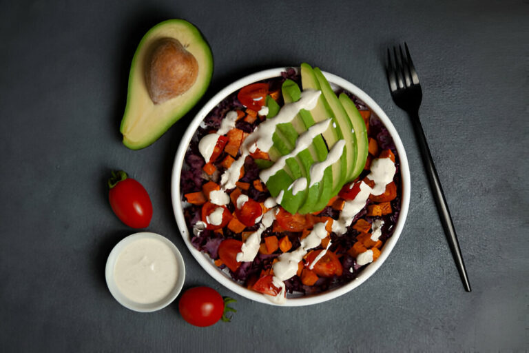 A colorful bowl of salad featuring avocado, tomatoes, and sweet potatoes, accompanied by a small bowl of dressing.