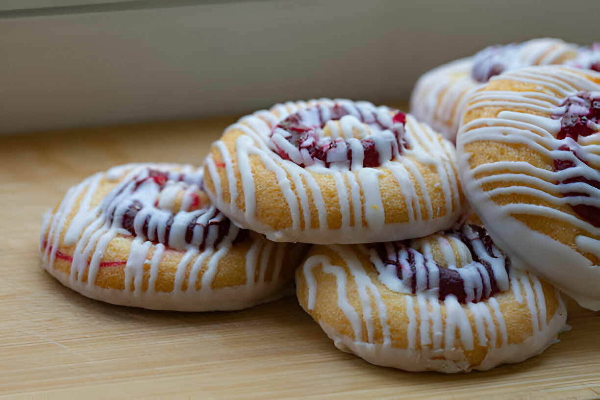 A close-up of delicious cookies topped with cherry filling and drizzled white icing, displayed on a wooden surface.