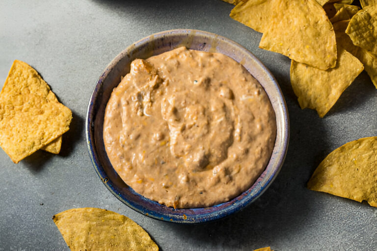 A bowl of creamy dip with crispy tortilla chips scattered around on a gray surface.