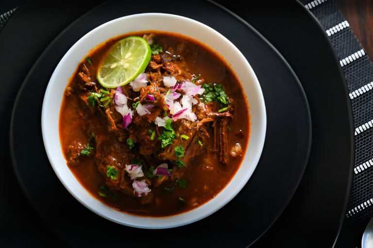 A delicious bowl of beef stew garnished with lime, onions, and herbs, served in a white bowl on a black plate.