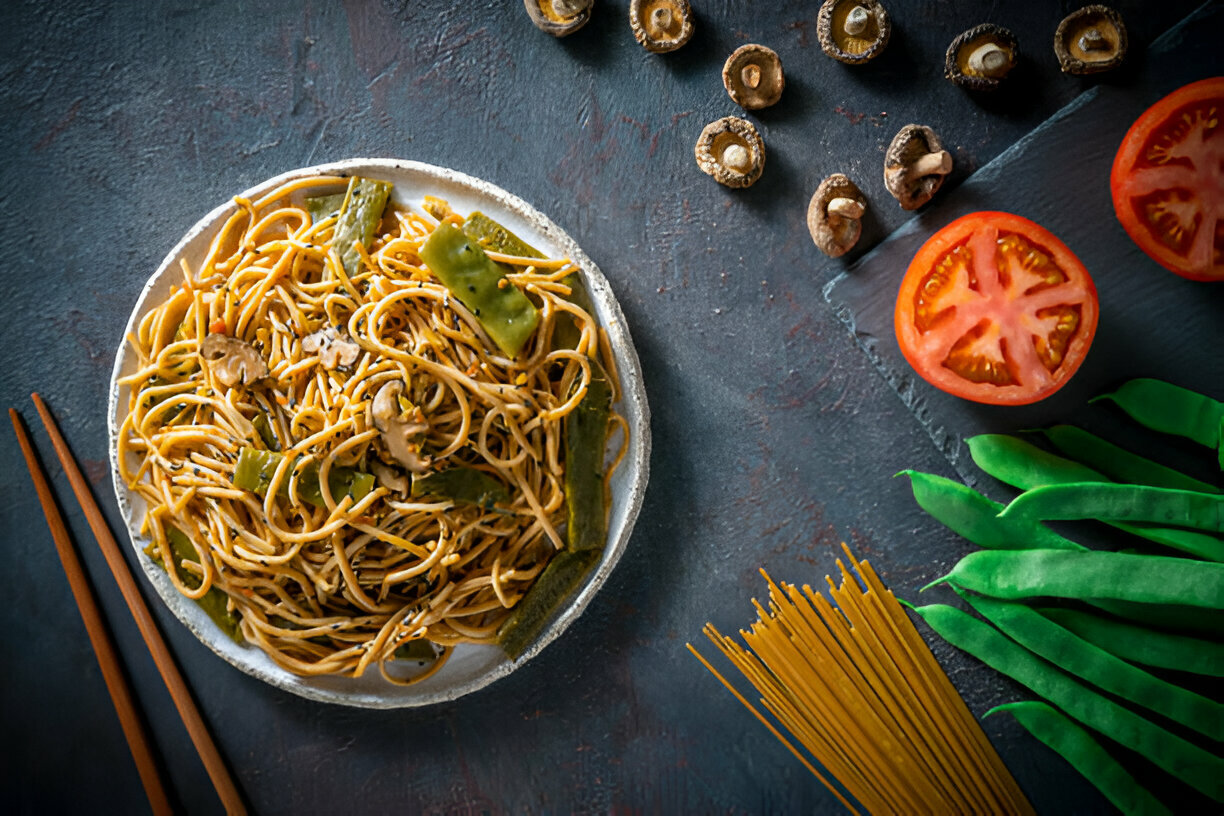 A plate of spaghetti topped with green vegetables and mushrooms, surrounded by fresh ingredients including sliced tomatoes, green beans, and dried mushrooms on a dark textured surface.