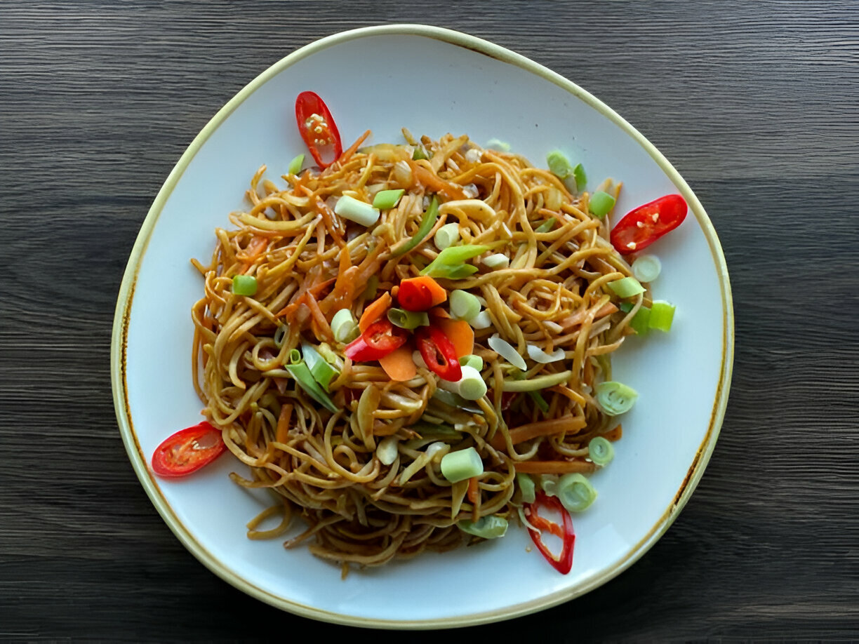 A plate of stir-fried noodles garnished with sliced green onions, red chilies, and vegetables.