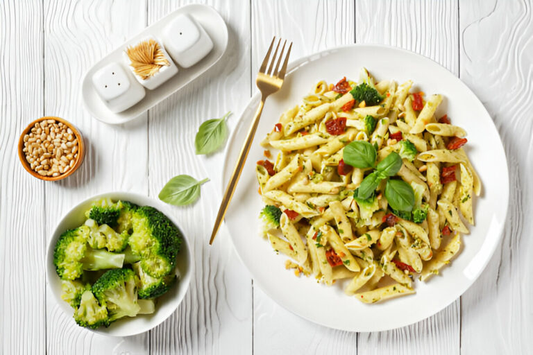 A delicious plate of pasta with basil and sun-dried tomatoes, accompanied by a side of steamed broccoli and pine nuts on a wooden table.